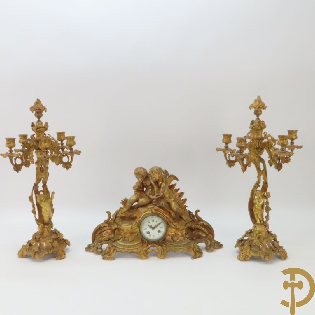 Bronzen schouwklok bezet met 2 cupido's, omgeven door accanthusranken en bladranken + paar bronzen vijfarmige kandelaars met centraal bloemenstuk en cupido's