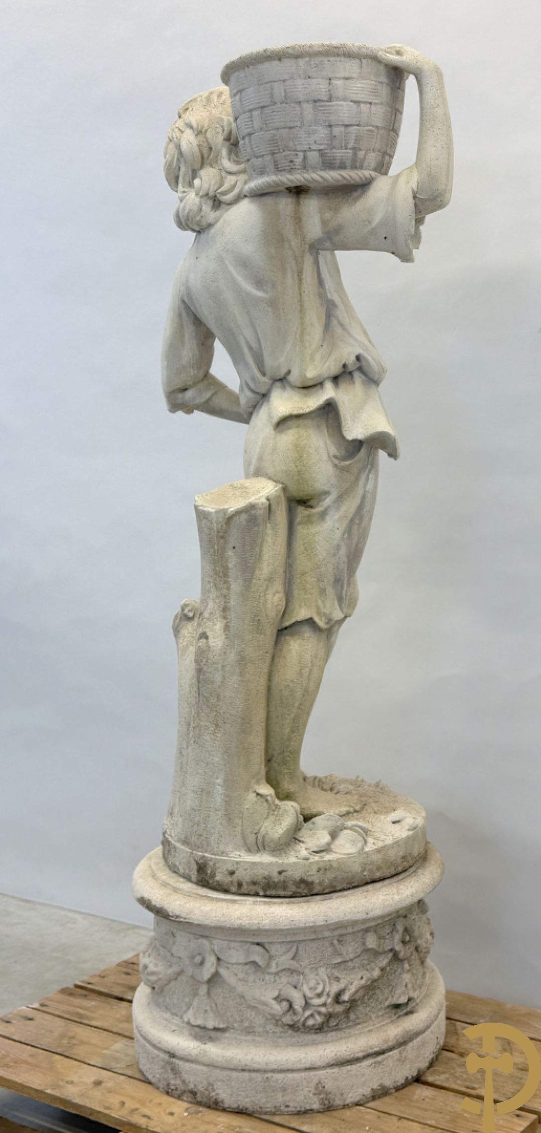 Stenen tuinbeeld van jongeman met mand in de hand op sokkel met guirlandes