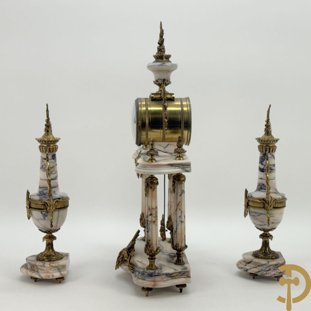 Driedelige wit marmeren garnituur bestaande uit vierzuilenpendule met bloemen en troffee geflankeerd door paar cassolettes met ramskoppen bezet