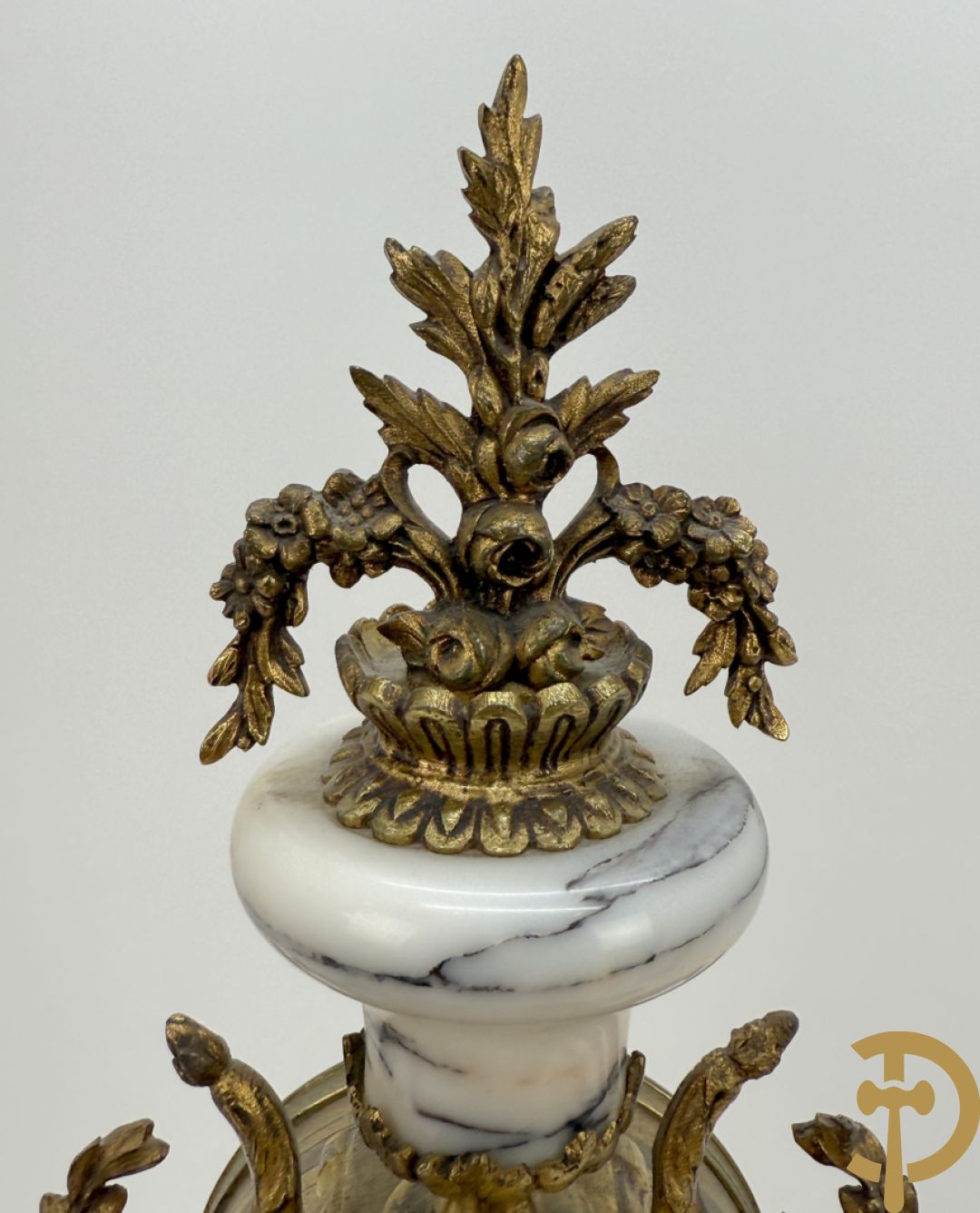 Driedelige wit marmeren garnituur bestaande uit vierzuilenpendule met bloemen en troffee geflankeerd door paar cassolettes met ramskoppen bezet