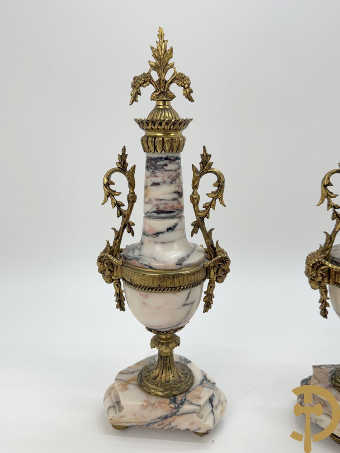 Driedelige wit marmeren garnituur bestaande uit vierzuilenpendule met bloemen en troffee geflankeerd door paar cassolettes met ramskoppen bezet