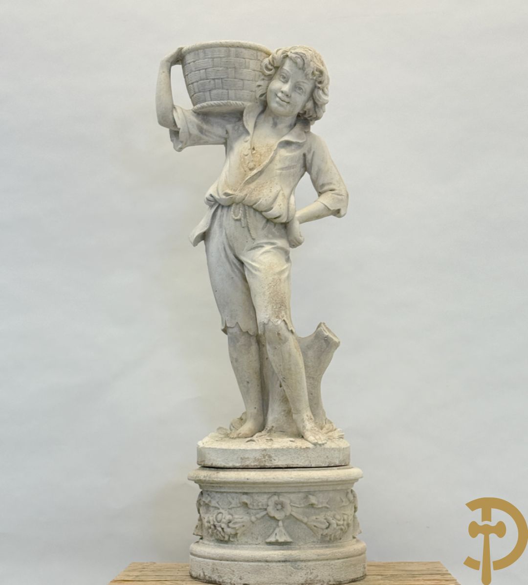 Stenen tuinbeeld van jongeman met mand in de hand op sokkel met guirlandes