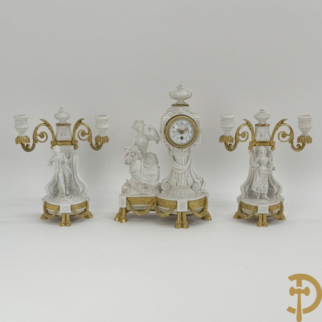 Driedelige porseleinen garnituur met bronsbeslag bestaande uit pendule met dame en amfoor geflankeerd door paar kandelaars bezet met personages