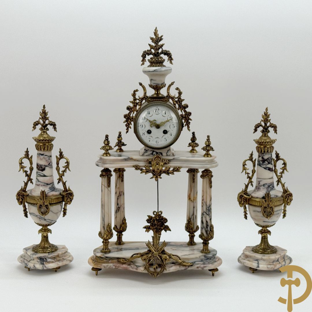 Driedelige wit marmeren garnituur bestaande uit vierzuilenpendule met bloemen en troffee geflankeerd door paar cassolettes met ramskoppen bezet