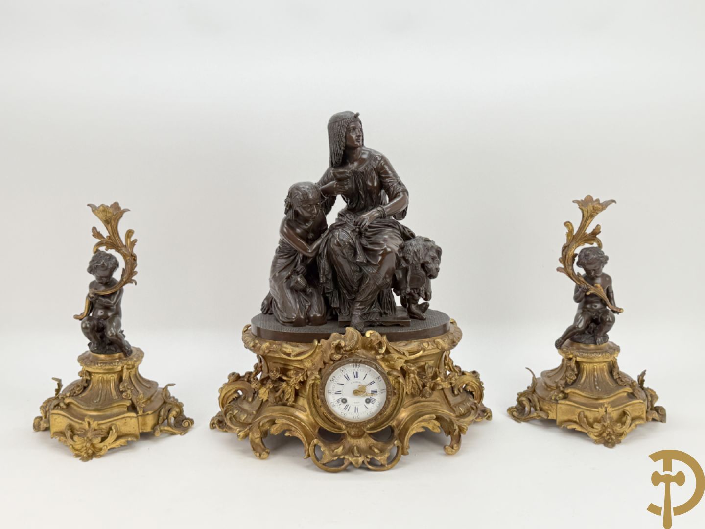 Driedelige bronzen garnituur bestaande uit middenstuk met bronzen vuurvergulde klok, Daubrée gemerkt à Nancy, bovenaan bekroond met Cleopatra met drinkbeker - getekend van Cumberworth sculpteur - geflankeerd door paar cupido