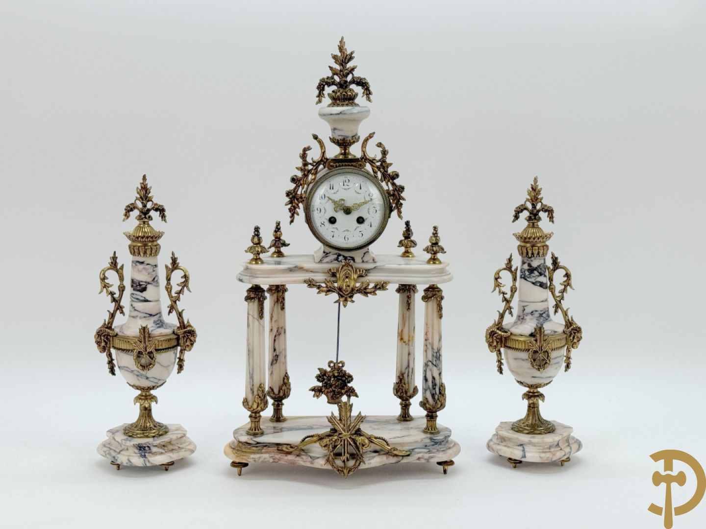 Driedelige wit marmeren garnituur bestaande uit vierzuilenpendule met bloemen en troffee geflankeerd door paar cassolettes met ramskoppen bezet