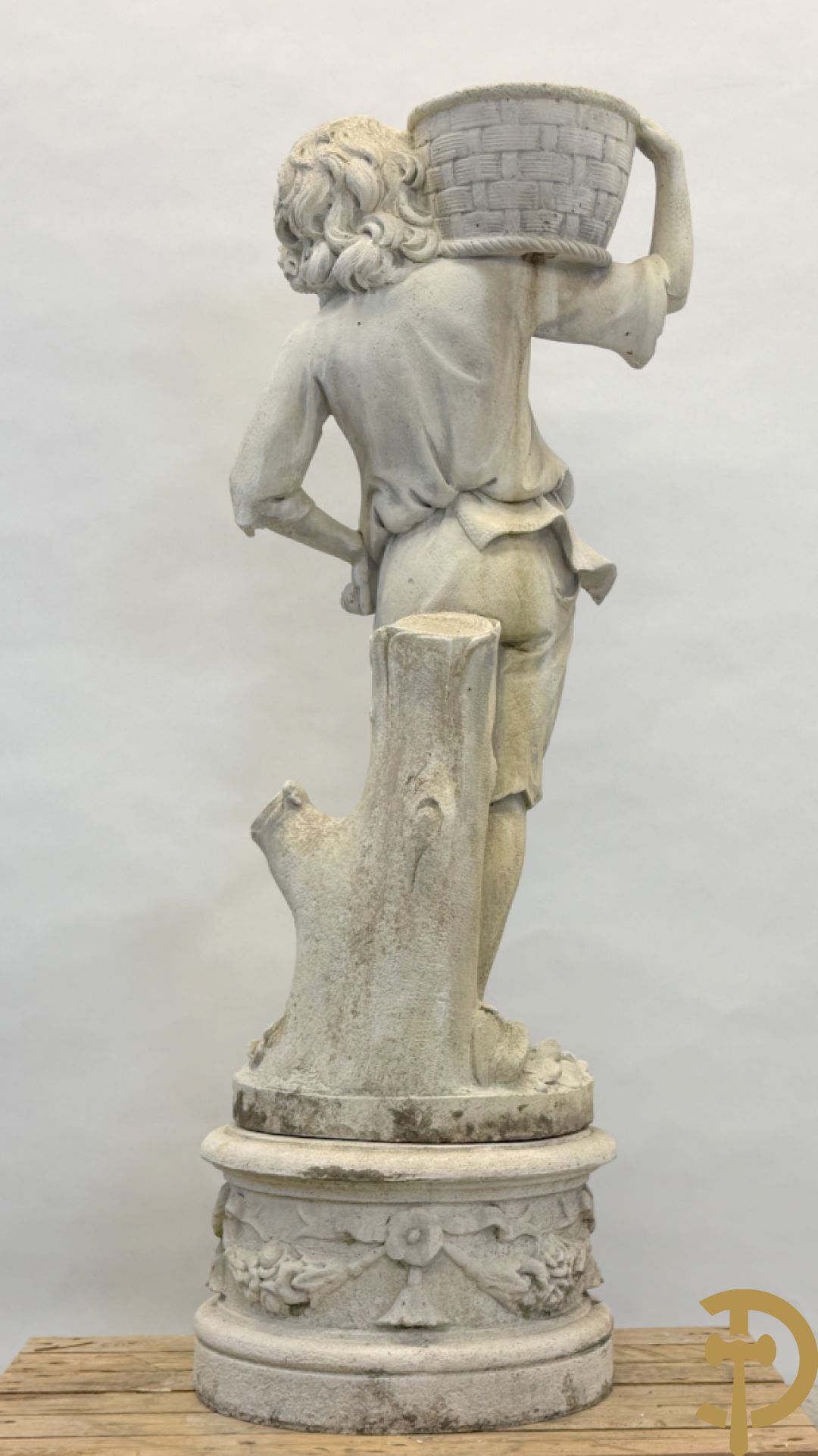 Stenen tuinbeeld van jongeman met mand in de hand op sokkel met guirlandes