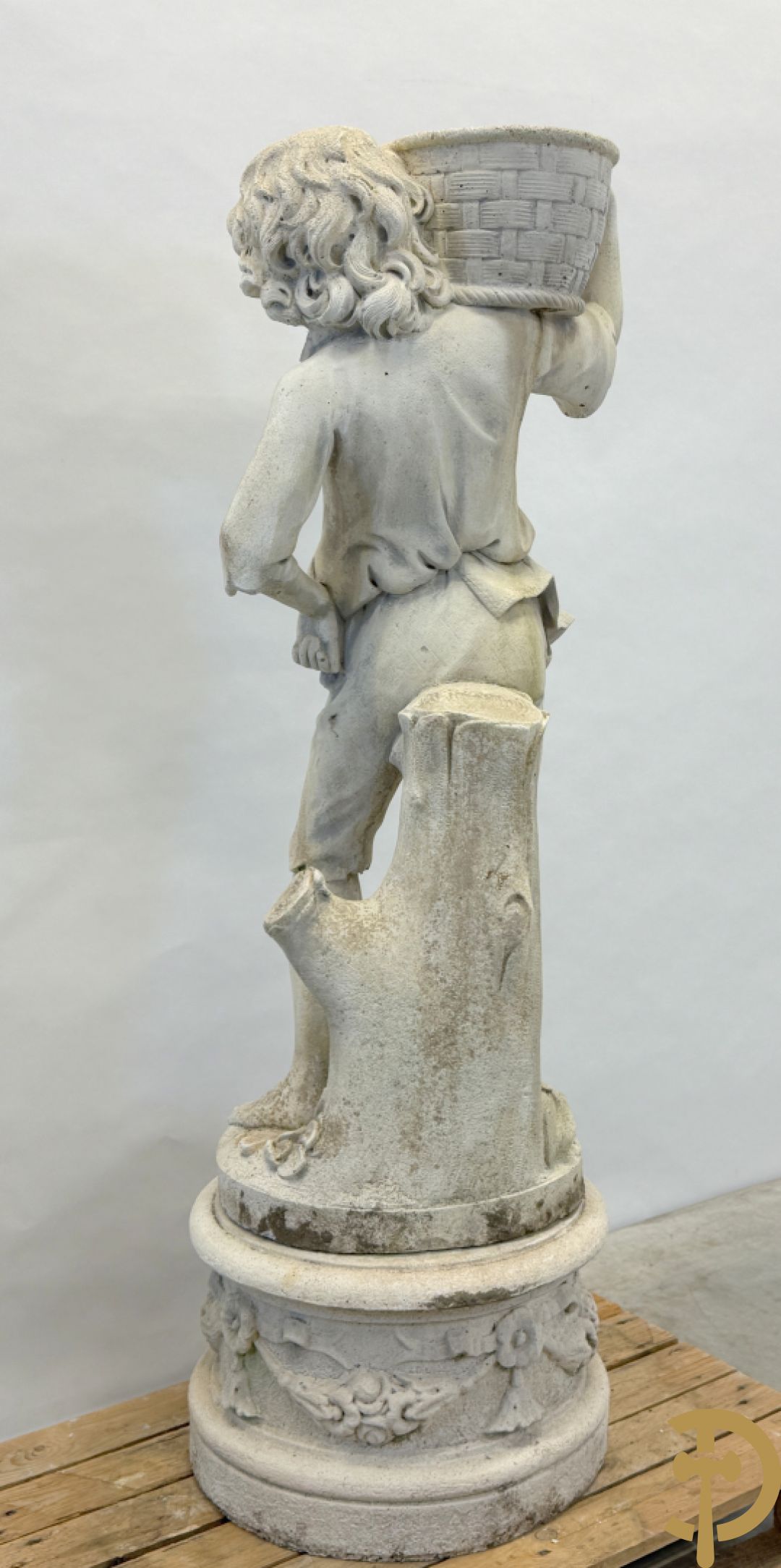 Stenen tuinbeeld van jongeman met mand in de hand op sokkel met guirlandes