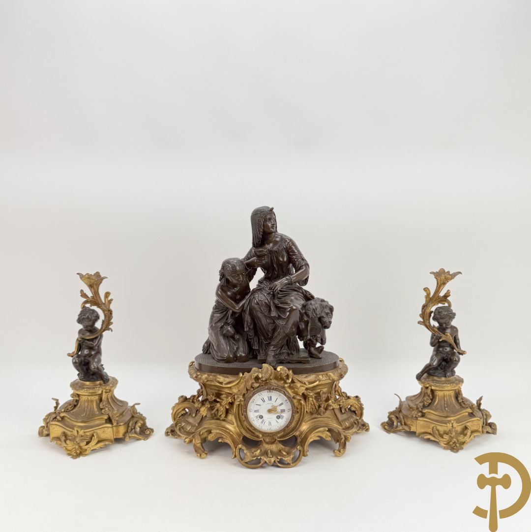 Driedelige bronzen garnituur bestaande uit middenstuk met bronzen vuurvergulde klok, Daubrée gemerkt à Nancy, bovenaan bekroond met Cleopatra met drinkbeker - getekend van Cumberworth sculpteur - geflankeerd door paar cupido
