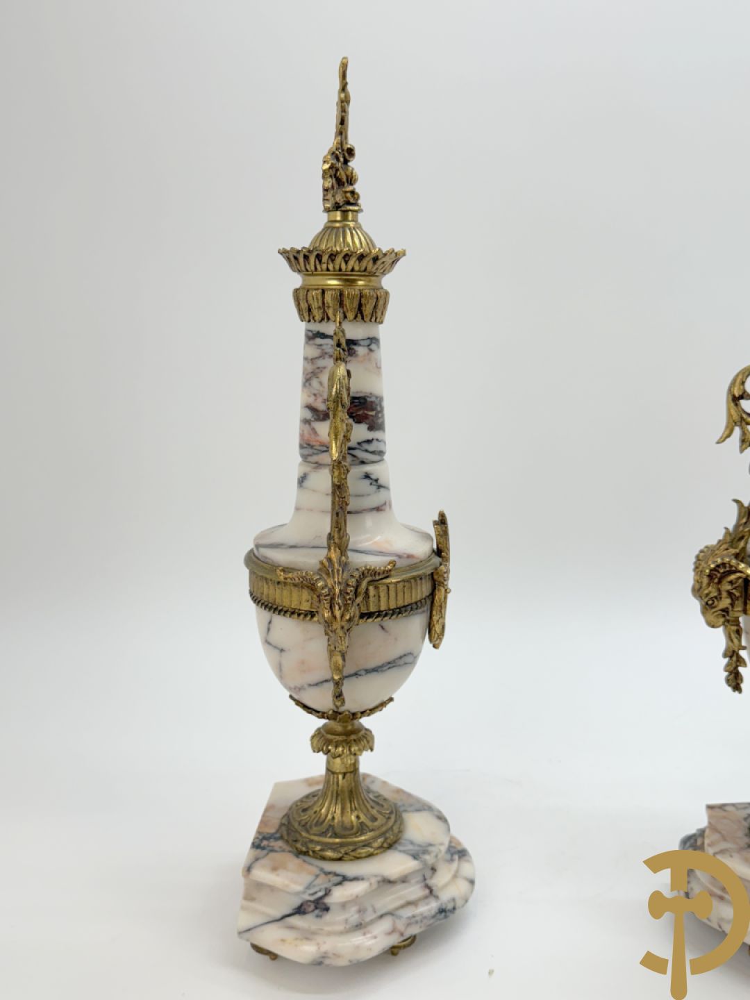 Driedelige wit marmeren garnituur bestaande uit vierzuilenpendule met bloemen en troffee geflankeerd door paar cassolettes met ramskoppen bezet