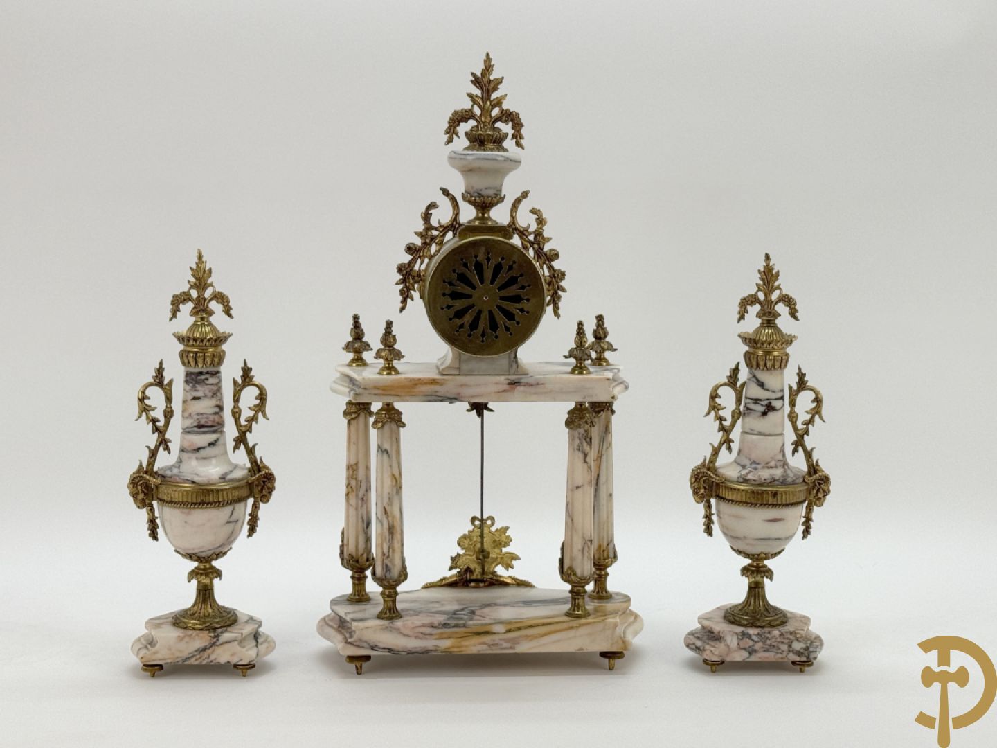 Driedelige wit marmeren garnituur bestaande uit vierzuilenpendule met bloemen en troffee geflankeerd door paar cassolettes met ramskoppen bezet