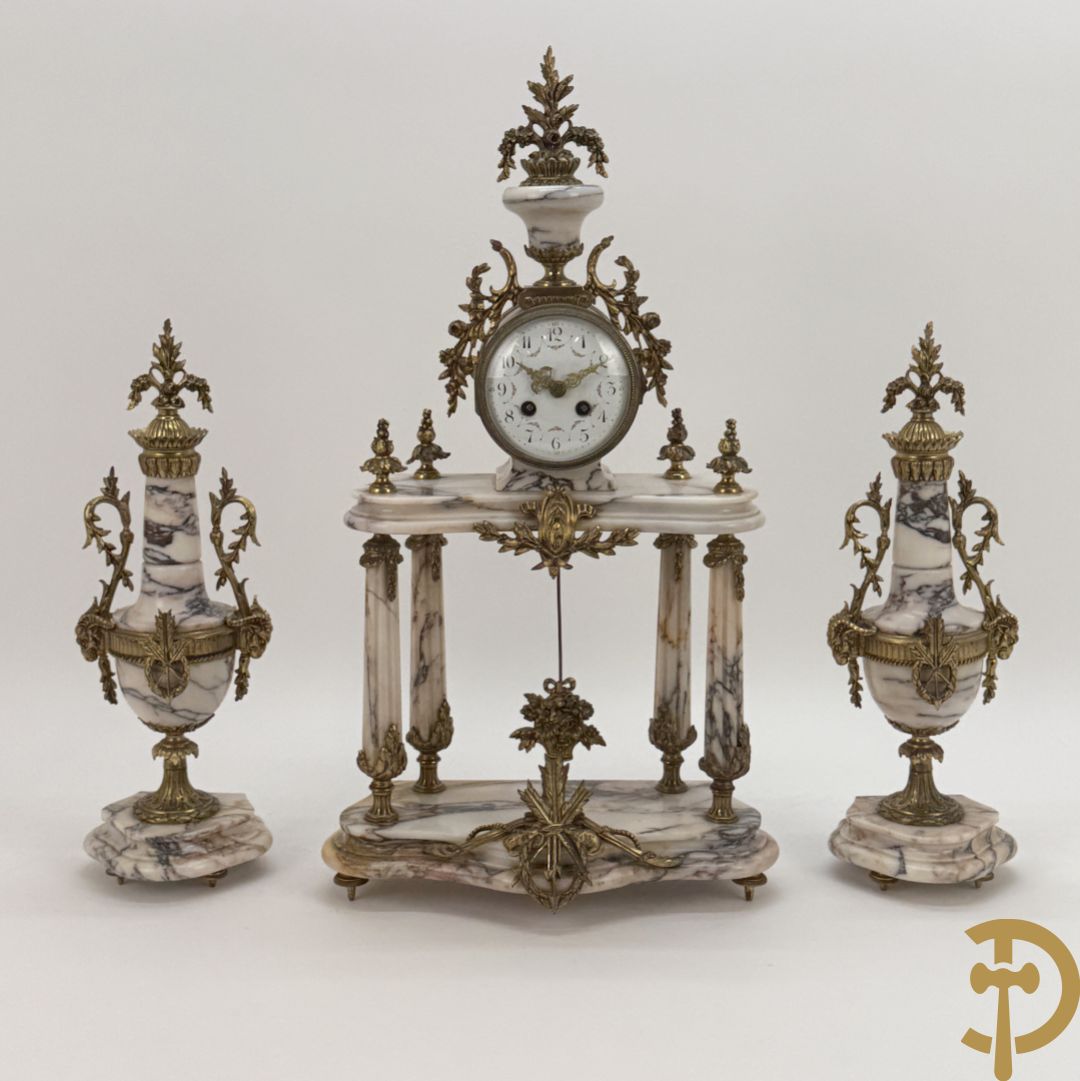 Driedelige wit marmeren garnituur bestaande uit vierzuilenpendule met bloemen en troffee geflankeerd door paar cassolettes met ramskoppen bezet