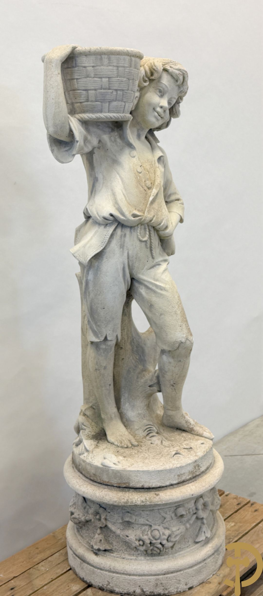 Stenen tuinbeeld van jongeman met mand in de hand op sokkel met guirlandes