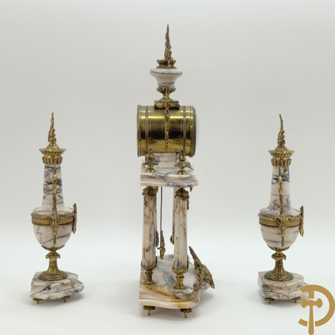 Driedelige wit marmeren garnituur bestaande uit vierzuilenpendule met bloemen en troffee geflankeerd door paar cassolettes met ramskoppen bezet