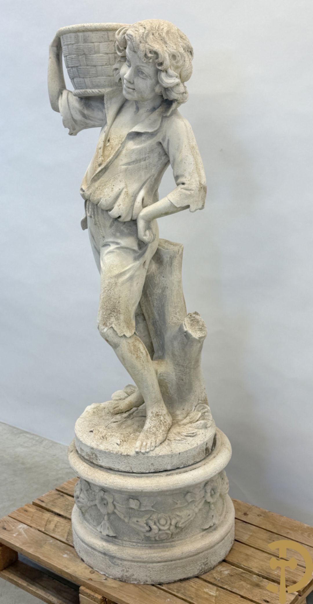 Stenen tuinbeeld van jongeman met mand in de hand op sokkel met guirlandes