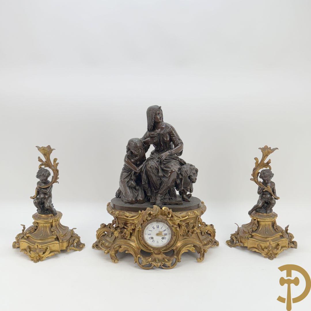Driedelige bronzen garnituur bestaande uit middenstuk met bronzen vuurvergulde klok, Daubrée gemerkt à Nancy, bovenaan bekroond met Cleopatra met drinkbeker - getekend van Cumberworth sculpteur - geflankeerd door paar cupido's (zonder kandelaars)