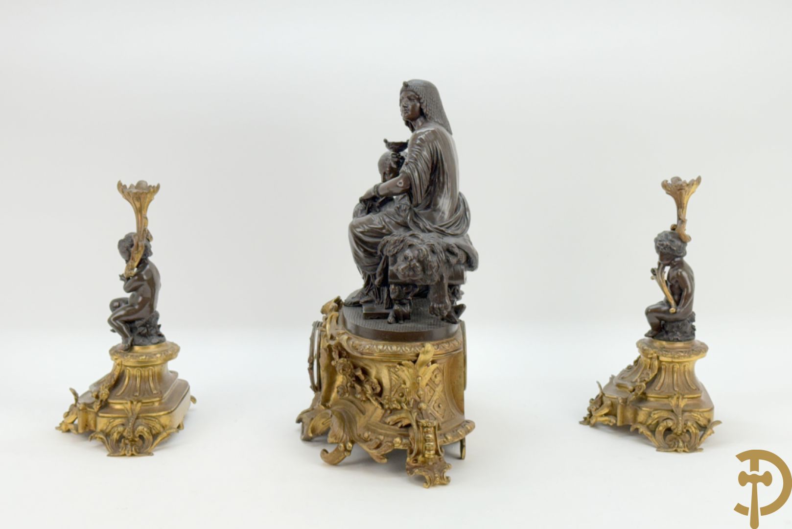 Driedelige bronzen garnituur bestaande uit middenstuk met bronzen vuurvergulde klok, Daubrée gemerkt à Nancy, bovenaan bekroond met Cleopatra met drinkbeker - getekend van Cumberworth sculpteur - geflankeerd door paar cupido