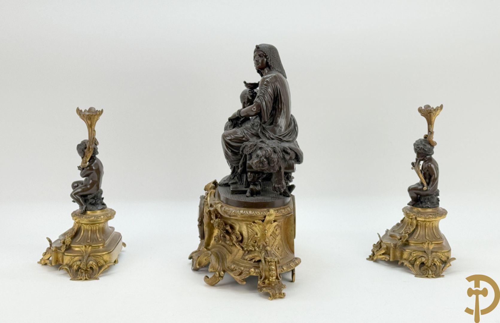 Driedelige bronzen garnituur bestaande uit middenstuk met bronzen vuurvergulde klok, Daubrée gemerkt à Nancy, bovenaan bekroond met Cleopatra met drinkbeker - getekend van Cumberworth sculpteur - geflankeerd door paar cupido