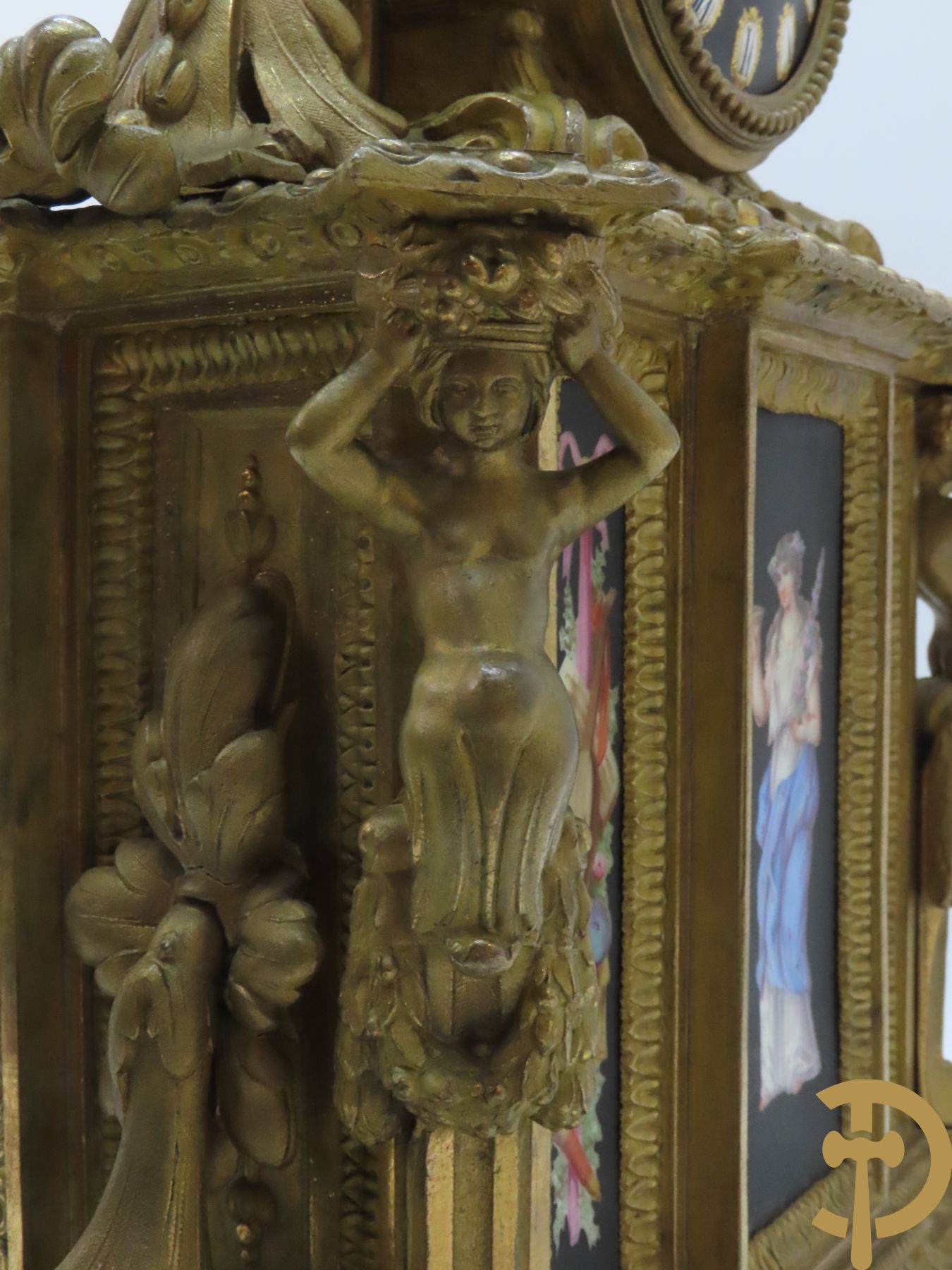 Vergulde pendule in zamak, wijzerplaat bezet met porseleinen plakkettes - bovenaan met vogelpaar, zijdelings met kariathyden houdende een bloementroffee - onder glazen stolp