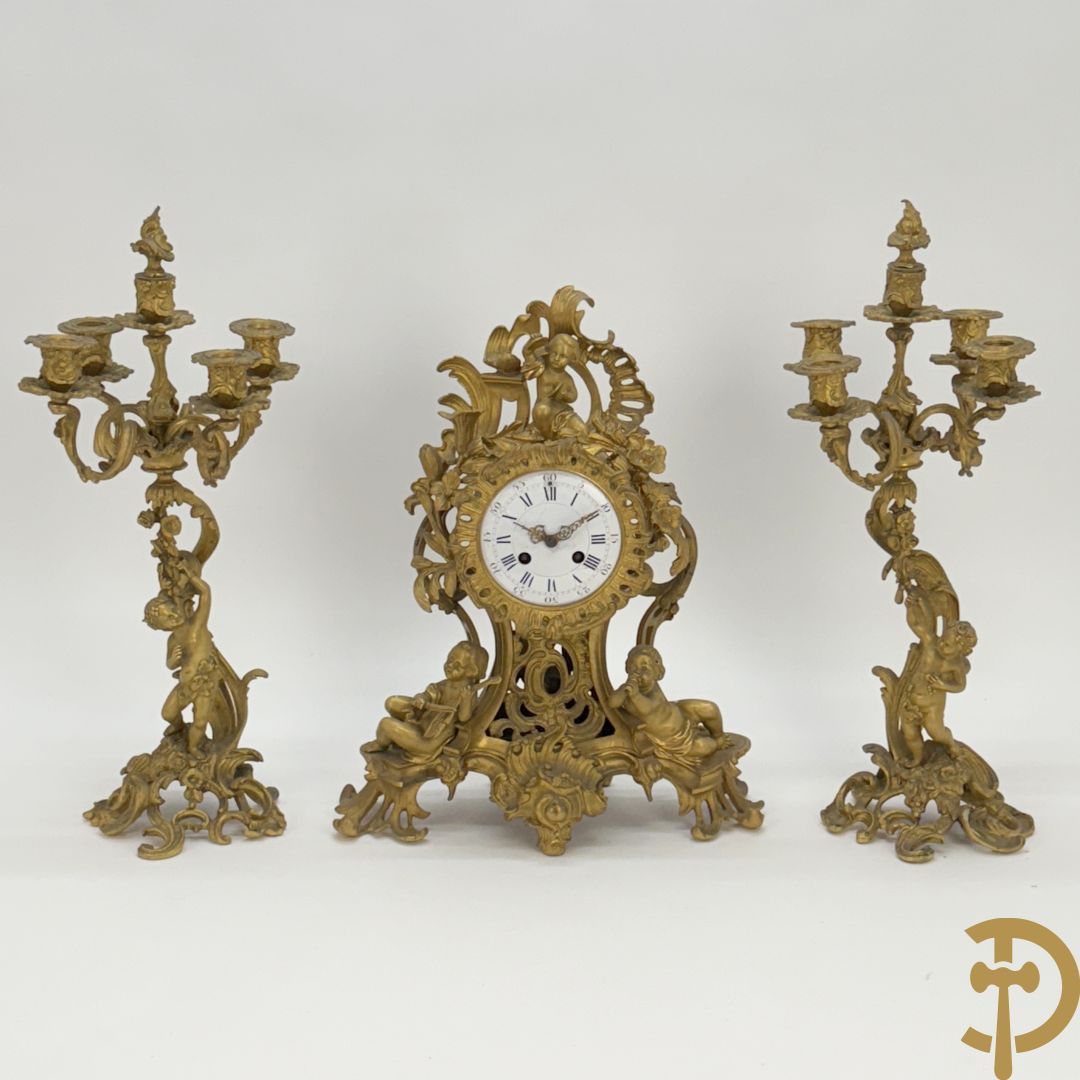 Driedelige vuurvergulde bronzen garnituur, Louis XV stijl, bestaande uit middenstuk met klok omgeven door musicerende cupido's en bloemen - geflankeerd door paar vijfarmige kandelaars