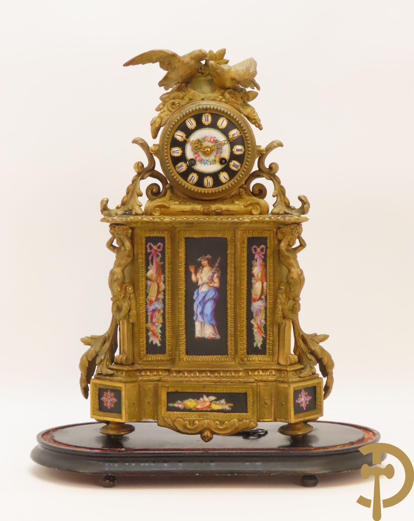 Vergulde pendule in zamak, wijzerplaat bezet met porseleinen plakkettes - bovenaan met vogelpaar, zijdelings met kariathyden houdende een bloementroffee - onder glazen stolp