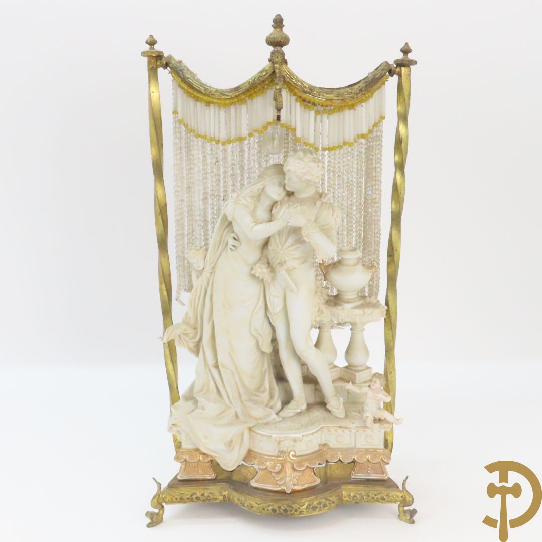 Lieftallig biscuit van romantisch koppel bij amfoor met kleine neergezeten cupido onder koperen baldakijn met kristallen belletjes