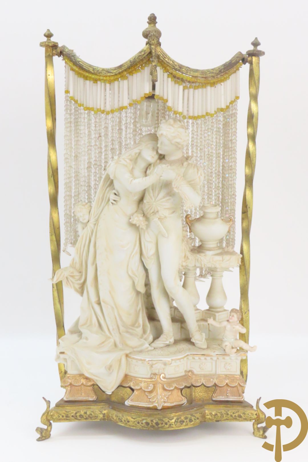 Lieftallig biscuit van romantisch koppel bij amfoor met kleine neergezeten cupido onder koperen baldakijn met kristallen belletjes