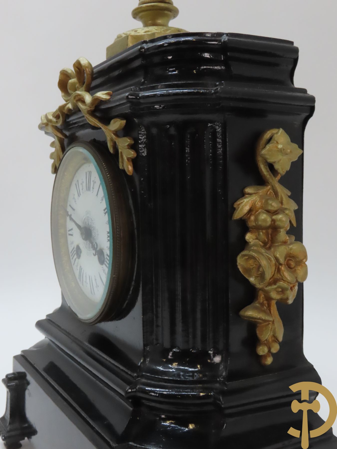 Gepatineerde gietijzeren pendule met bloemenmotieven en Louis XVI strik, bovenaan bekroond met ornament